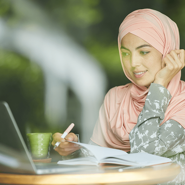 A young girl sporting a smile as she looks at a computer intently while taking notes. Almost as if she is attending a webinar.